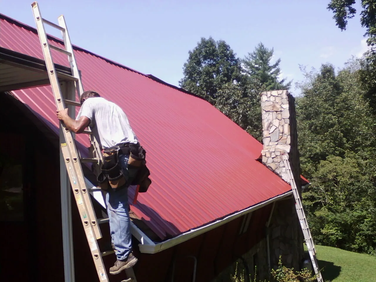 Roofer on a ladder beside a red metal roof and stone chimney — Soffit & Fascia Repair by Roofing Brothers in Morton